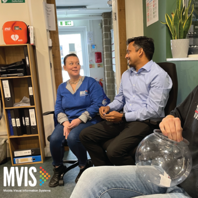 A man and a woman sat, 2 MVIS Staff, on a chair laughing together with the MVIS logo in the bottom left corner.